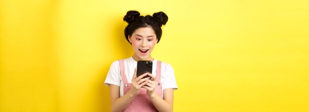 Jolie fille asiatique regardant excitée par un message de lecture d'écran sur le téléphone et souriante heureuse debout dans s