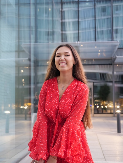 Jolie fille asiatique souriante en robe rouge sourit à côté des façades en verre des bâtiments.