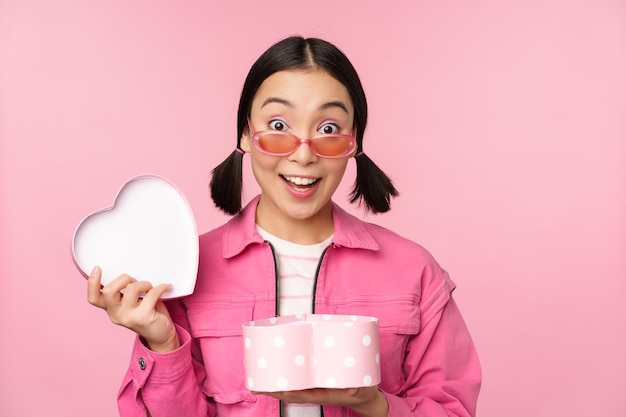 Jolie fille asiatique vous donnant un cadeau dans une boîte en forme de coeur embrassant et souriant concept de vacances et de célébration debout sur fond rose