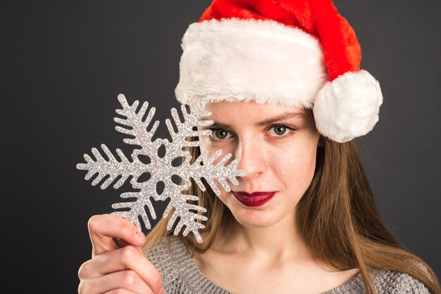 Jolie fille au chapeau de père noël tient un gros flocon de neige argenté près du visage