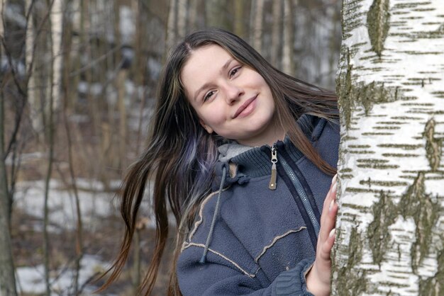 Une jolie fille au printemps dans la nature regarde derrière un bouleau.