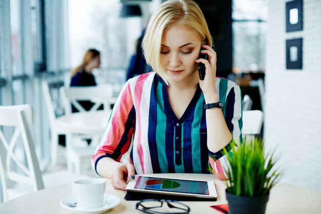 Jolie fille aux cheveux clairs portant une chemise colorée assis dans un café avec tablette, téléphone portable et tasse de café, portrait, concept indépendant, achats en ligne.