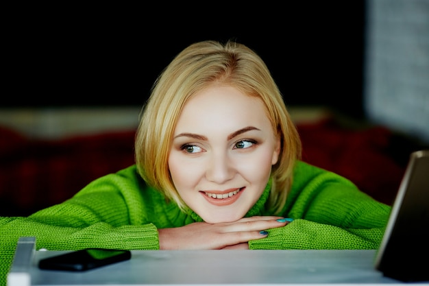 Jolie fille aux cheveux clairs portant un pull vert assis dans un café avec téléphone portable et tablette, concept indépendant, achats en ligne, portrait.