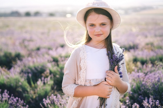 Jolie fille aux cheveux longs dans une robe en lin et un chapeau avec un bouquet de lavande debout dans un champ de lavande