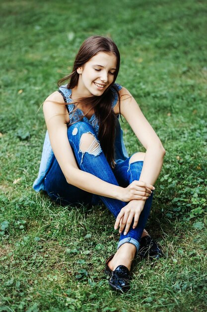 Jolie fille aux cheveux longs et jeans déchirés souriant avec les yeux fermés assis sur l'herbe dans le parc en plein air