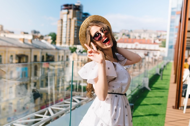 Jolie fille aux cheveux longs en lunettes de soleil pose pour la caméra sur la terrasse. Elle porte une robe blanche aux épaules nues, du rouge à lèvres et un chapeau. Elle sourit à la caméra.