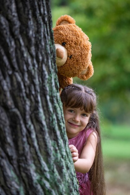 Une jolie fille aux cheveux longs regarde derrière un grand arbre. Au-dessus d'elle se trouve un gros ours en peluche.