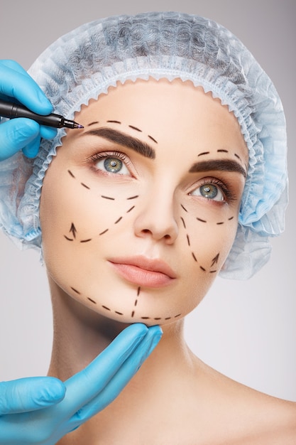 Jolie fille aux sourcils noirs portant un chapeau médical bleu à fond de studio, les mains du médecin portant des gants bleus dessinant des lignes de perforation sur le visage, concept de chirurgie plastique.