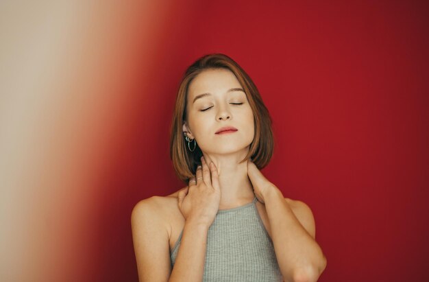 Jolie fille aux yeux fermés et des vêtements décontractés se dresse sur un fond rouge et pose à la caméra avec un sourirePortrait créatif d'une jolie fille appréciant les yeux fermés sur un fond coloré