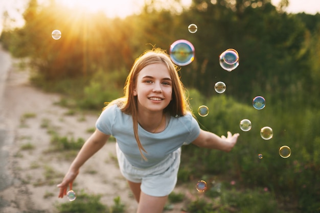 Jolie fille avec un beau sourire attrape des bulles de savon sur le fond d'un beau coucher de soleil