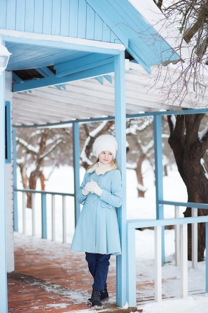 Jolie fille blonde dans un manteau bleu et un chapeau de fourrure blanche en hiver. Temps neigeux. La jeune fille sur le porche de la maison. Maison d'hiver. Modèle posant dans la rue. Le concept de vacances d'hiver.