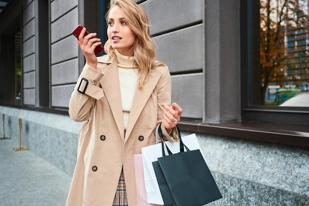 Jolie fille blonde décontractée en manteau beige avec des sacs à provisions enregistrant des messages vocaux sur un téléphone portable dans la rue de la ville