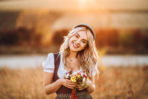 Jolie Fille Blonde En Dirndl, Debout à L'extérieur Dans Le Domaine, Tenant Le Bouquet D'un Champ De Fleurs.