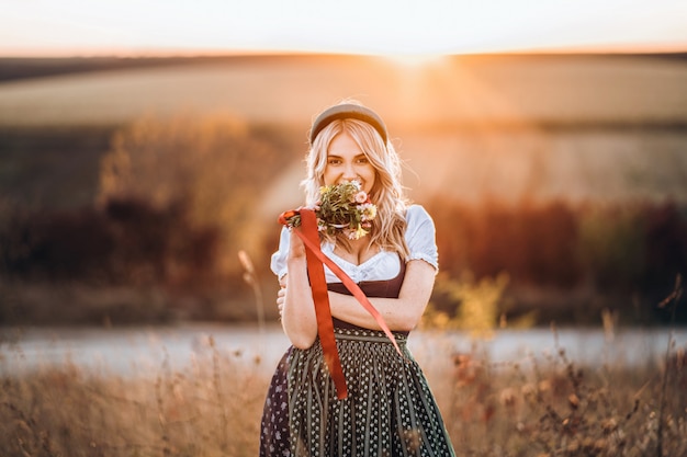 Jolie fille blonde en dirndl, debout à l'extérieur dans le domaine, tenant le bouquet d'un champ de fleurs.