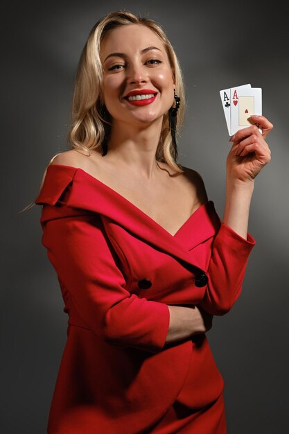 Jolie fille blonde à l'épaule nue, maquillage lumineux, en robe rouge et boucles d'oreilles noires. Elle sourit, montrant deux cartes à jouer, posant sur fond gris. Poker, casino. Gros plan, espace de copie