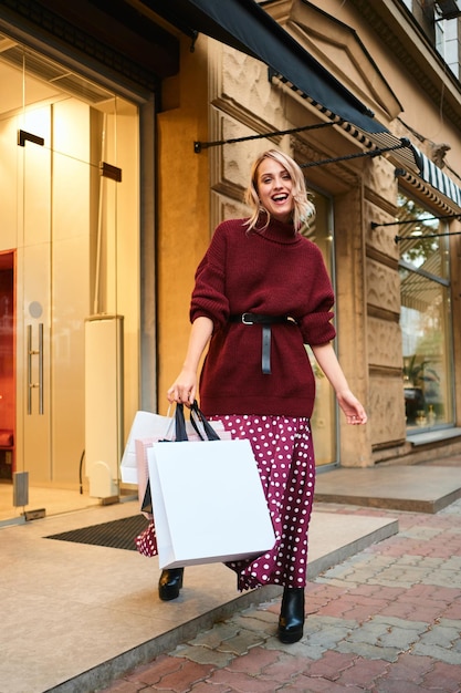 Jolie fille blonde joyeuse en pull tricoté avec des sacs à provisions regardant joyeusement à huis clos après le shopping en plein air