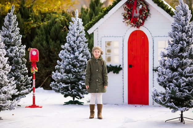 Jolie fille blonde près de la petite maison et des arbres enneigés. Nouvel An et Noël