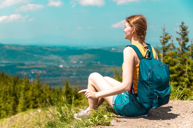 Jolie fille blonde avec un sac à dos et des lunettes est assis sur une montagne et profite des belles collines de la montagne par une journée ensoleillée.