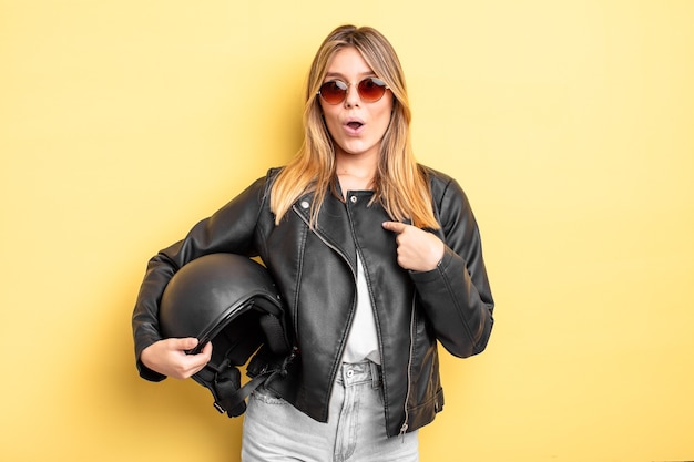 Jolie fille blonde semblant choquée et surprise avec la bouche grande ouverte, pointant vers soi. concept de casque de moto