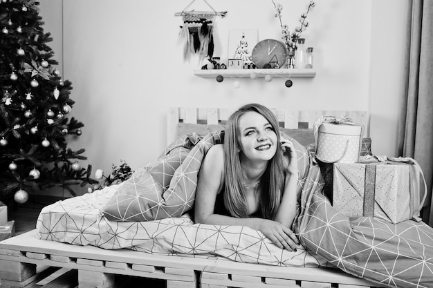 Jolie fille blonde sur soutien-gorge noir allongé sur le lit avec des coffrets cadeaux contre l'arbre du nouvel an au studio Joyeuses fêtes