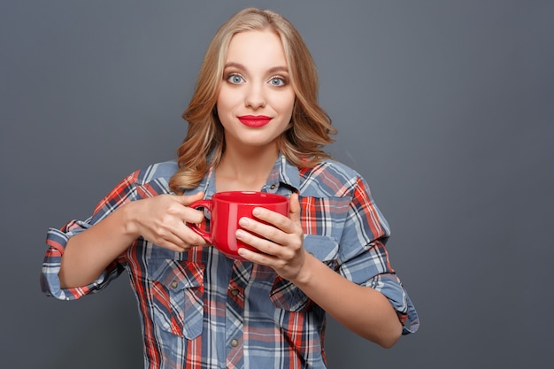 Jolie fille blonde tient une tasse rouge et regarde droit devant