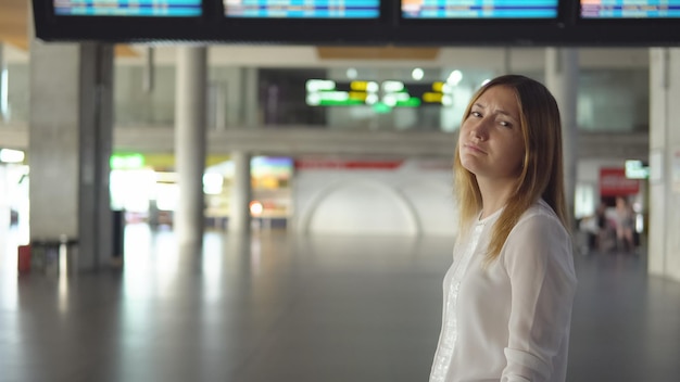 Jolie fille blonde en vêtements décontractés est triste dans un terminal d'aéroport vide et regarde dans la caméra