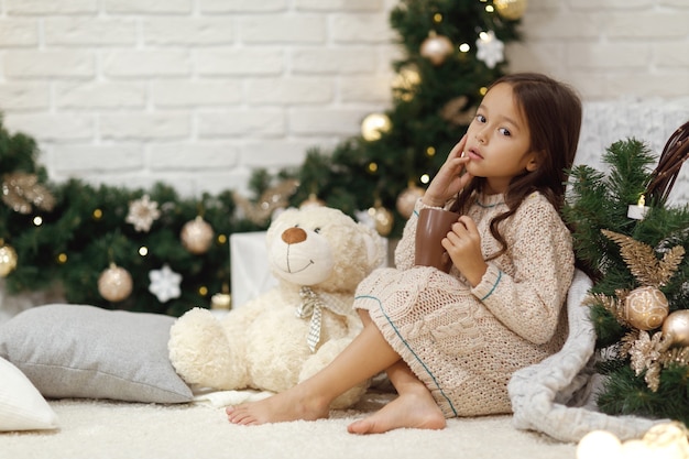 Jolie fille boit du chocolat chaud et joue avec un ours en peluche à la maison pour Noël