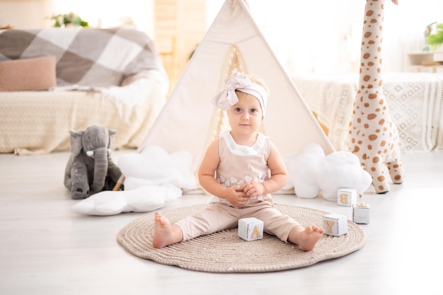 Une jolie fille en bonne santé est assise sur un tapis dans le salon lumineux de la maison sur le fond d'un wigwam et de jouets en peluche jouant avec des jouets éducatifs en bois textiles de maison