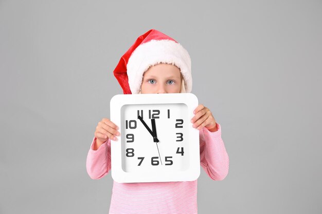 Jolie fille en bonnet de Noel avec horloge sur fond gris. Notion de compte à rebours de Noël