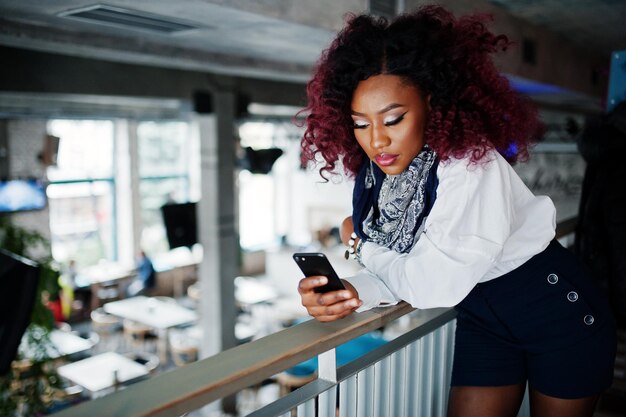 Jolie fille bouclée afro-américaine en blouse blanche regardant le téléphone au club