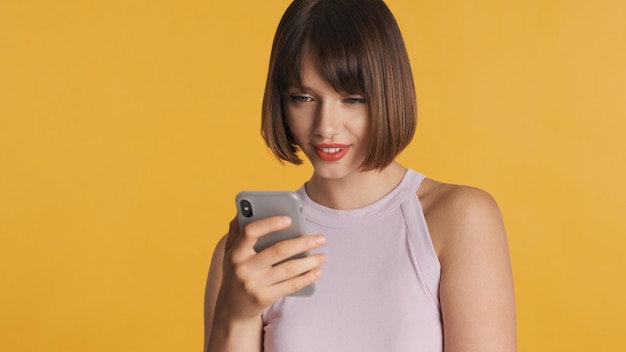 Jolie fille brune aux cheveux bob discutant avec des amis en ligne sur smartphone sur fond jaune Toujours en contact