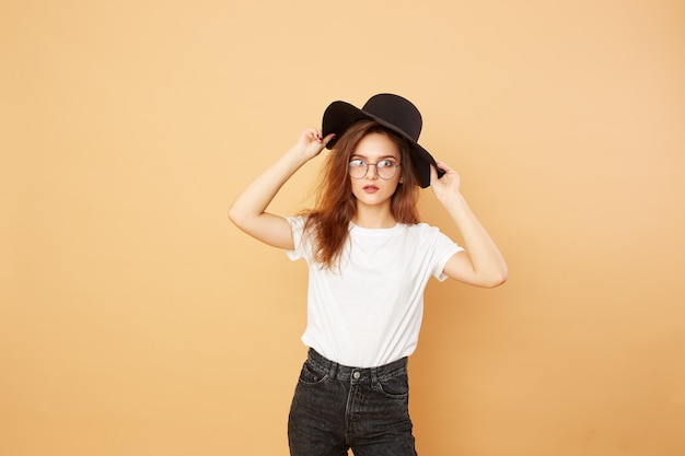 Jolie fille brune aux cheveux longs dans le chapeau noir sur la tête vêtue d'un t-shirt blanc et d'un jean noir pose sur le fond beige en studio.