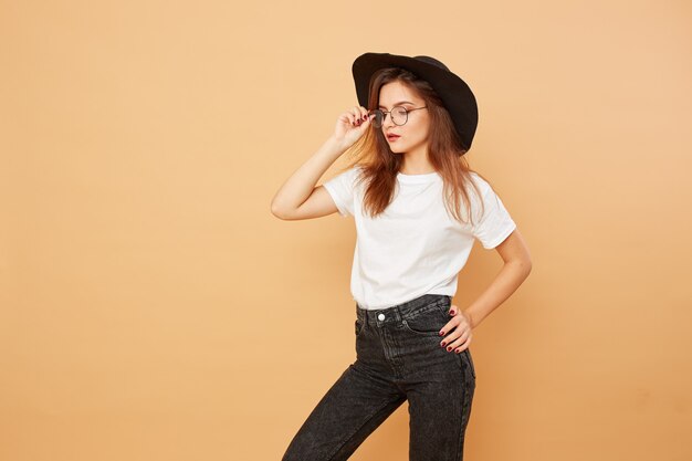 Jolie fille brune aux cheveux longs dans le chapeau noir sur la tête vêtue d'un t-shirt blanc et d'un jean noir pose sur le fond beige en studio.