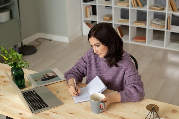 une jolie fille brune dans un pull lilas est assise à une table avec un ordinateur portable prenant des notes