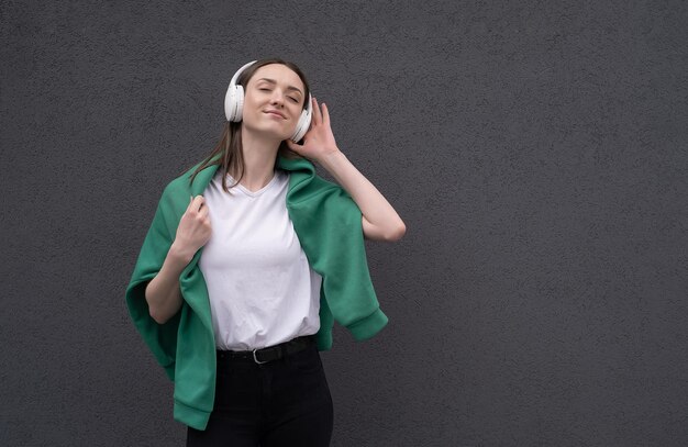 Jolie fille brune insouciante vêtue d'un pull gris écoutant de la musique sur un casque debout