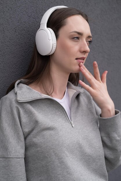 Jolie fille brune insouciante vêtue d'un pull gris écoutant de la musique sur un casque debout