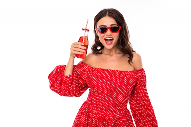 Jolie fille brune à lunettes de soleil et robe rouge avec un verre de cocktail sur un mur blanc