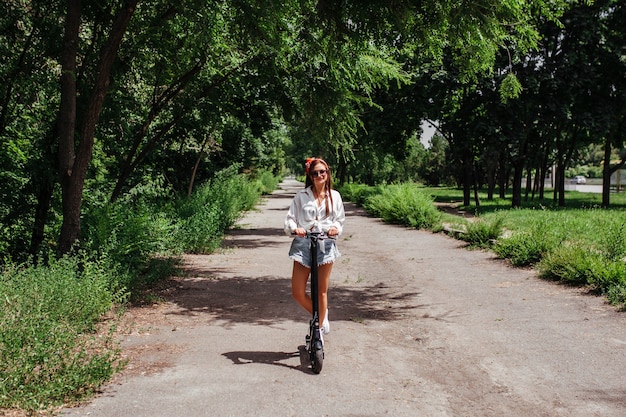 Jolie fille brune monte sur un scooter à électrodes dans le parc, elle porte une chemise blanche. concept de transport écologique et de location.