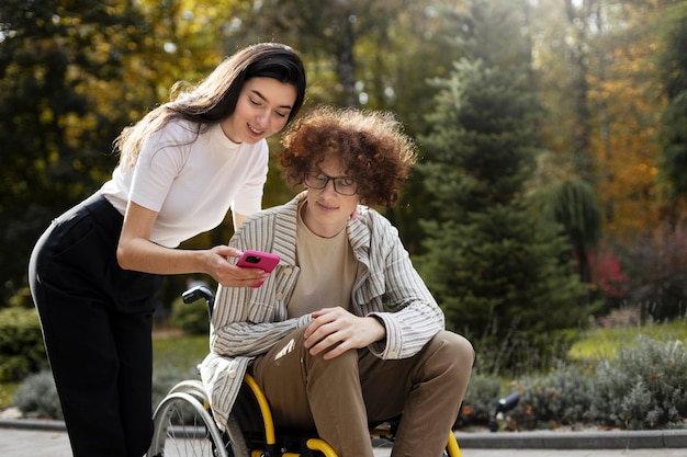 Jolie fille brune montre son téléphone à son ami handicapé Un jeune homme est assis dans un fauteuil roulant en plein air