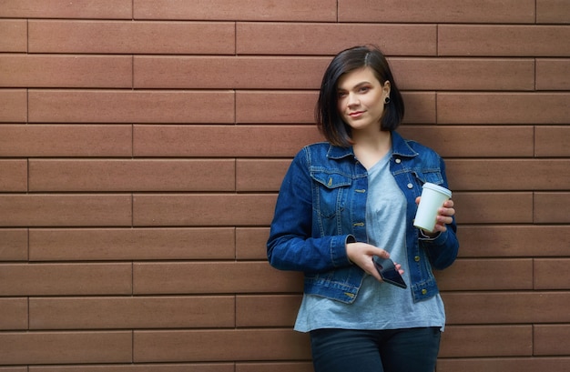 Jolie fille brune avec des tunnels dans les oreilles dans une veste en jean bleu avec une tasse de café et un smartphone debout devant le mur de briques.