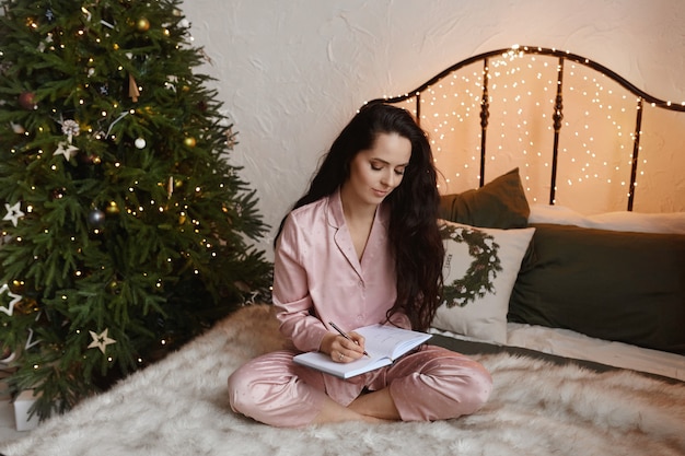 Jolie fille brune en vêtements de nuit confortables, assise sur le lit et écrivant les plans du nouvel an dans un cahier à côté de l'arbre de Noël.