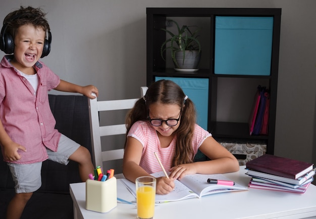 Une jolie fille caucasienne en colère à lunettes étudie à la maison, fait ses devoirs. Petit frère déranger. Chantez et criez. L'écriture. concept d'éducation à distance. Quarantaine. Retour au concept de l'école.