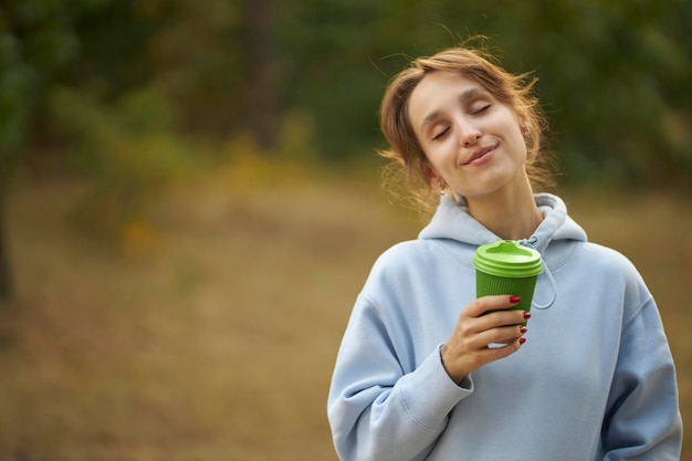 Jolie fille caucasienne dans un sweat à capuche bleu garde les yeux fermés, sourit doucement, tient du café à emporter.