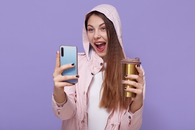 Jolie fille caucasienne faisant selfie sur fond lilas, excitée, gardant la bouche largement ouverte, appréciant une boisson chaude, portant un imperméable rose pâle.