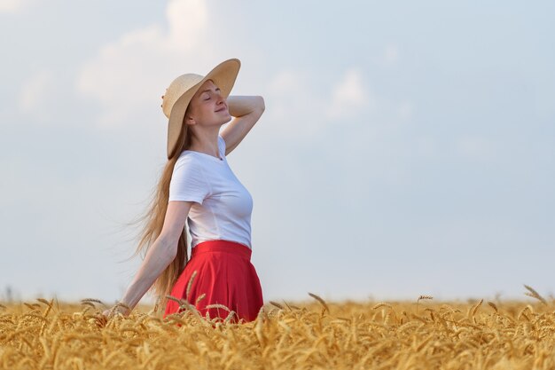 Jolie fille avec un chapeau se promène dans le champ de blé et profite d'une journée ensoleillée.