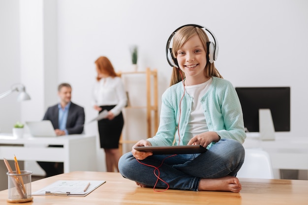 Jolie fille charmante intelligente assise sur la table et jouant avec son gadget pendant que son père travaille dur sur son projet