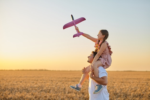 Jolie fille à cheval sur l'épaule du père et jouant avec un avion jouet contre le ciel dans un champ de blé