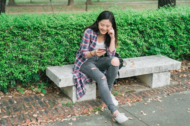 Jolie fille chinoise asiatique qui rit avec un smartphone passe du bon temps le week-end d'été. Portrait extérieur d'une jolie dame branchée assise sur un banc de parc et utilisant un téléphone portable s'amusant en souriant par sac à côté.