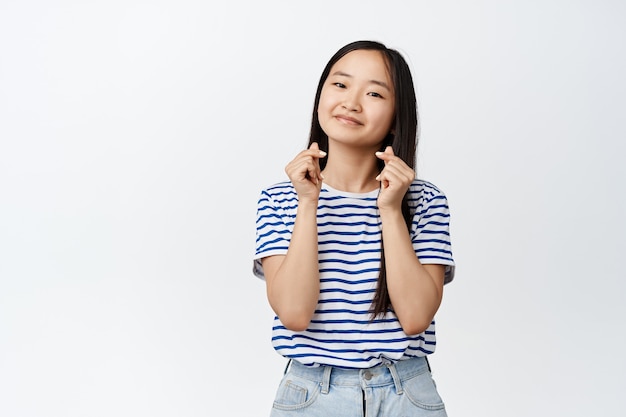 Une jolie fille chinoise montre un geste de coeur de doigt et une expression souriante et adorable du visage, debout en t-shirt sur blanc