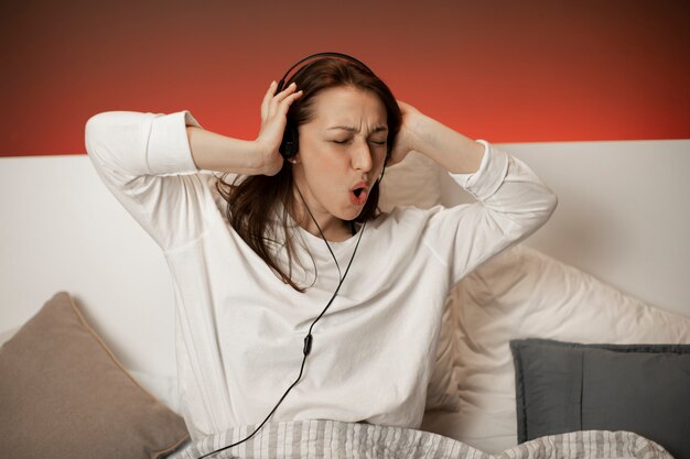 Photo jolie fille contenue en pyjama, écouter de la musique à l'aide d'écouteurs et danser sur le lit